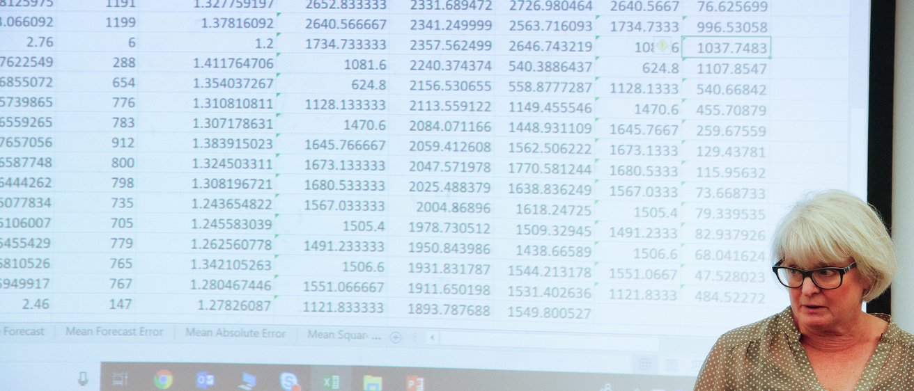 Instructor speaking in front of a business analytics class, with data on the screen behind her